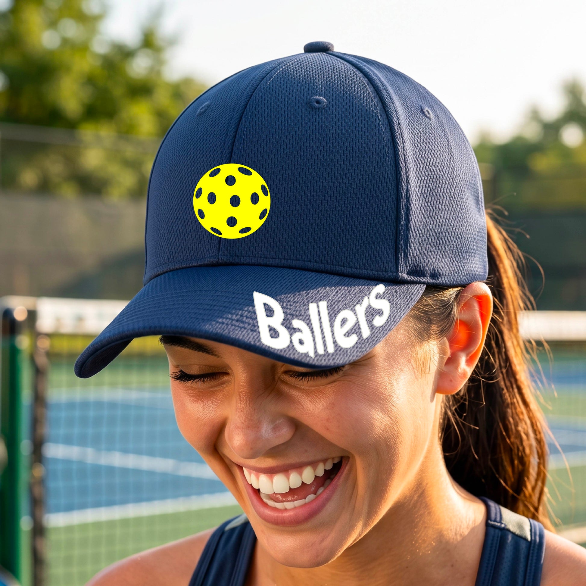 A person on a pickleball court wearing a navy blue baseball cap with a pickleball graphic on the front and the word 'Ballers' in white on the brim.