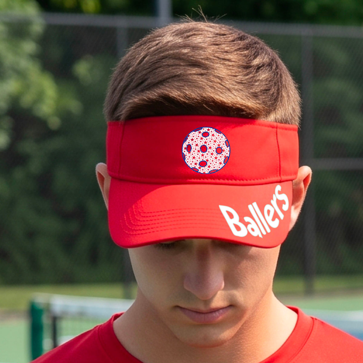 A person wearing a red visor with a yellow pickleball graphic and the word 'Ballers' in white,.