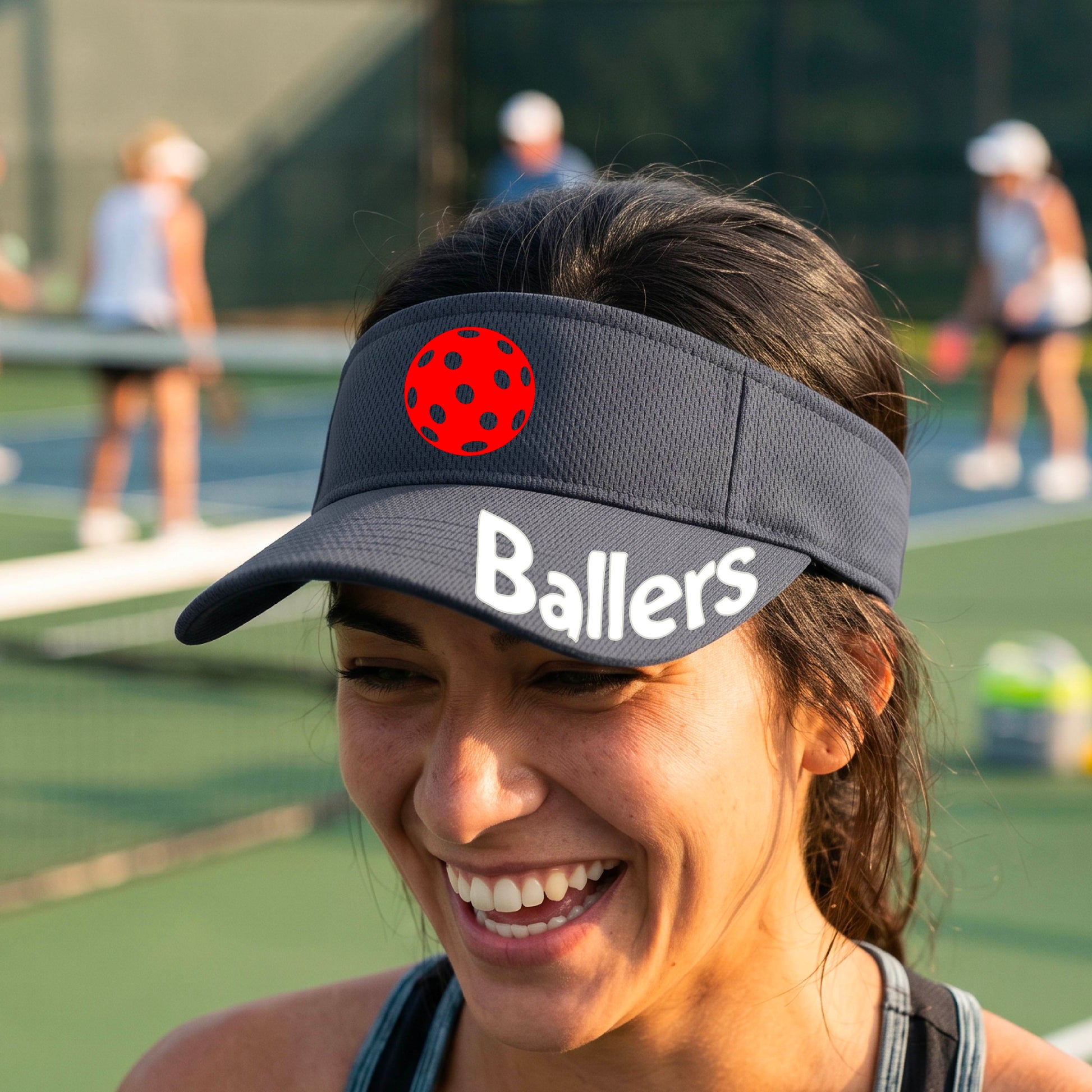 A person wearing a graphite gray visor with a yellow pickleball graphic and the word 'Ballers' in white,.