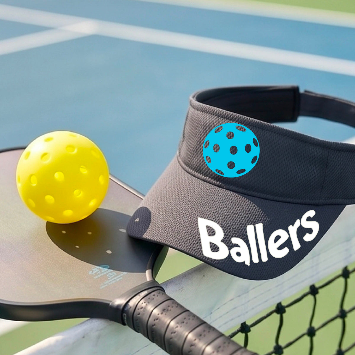 A close up of a graphite gray visor with pickleball graphic and the word 'Ballers' in white,.