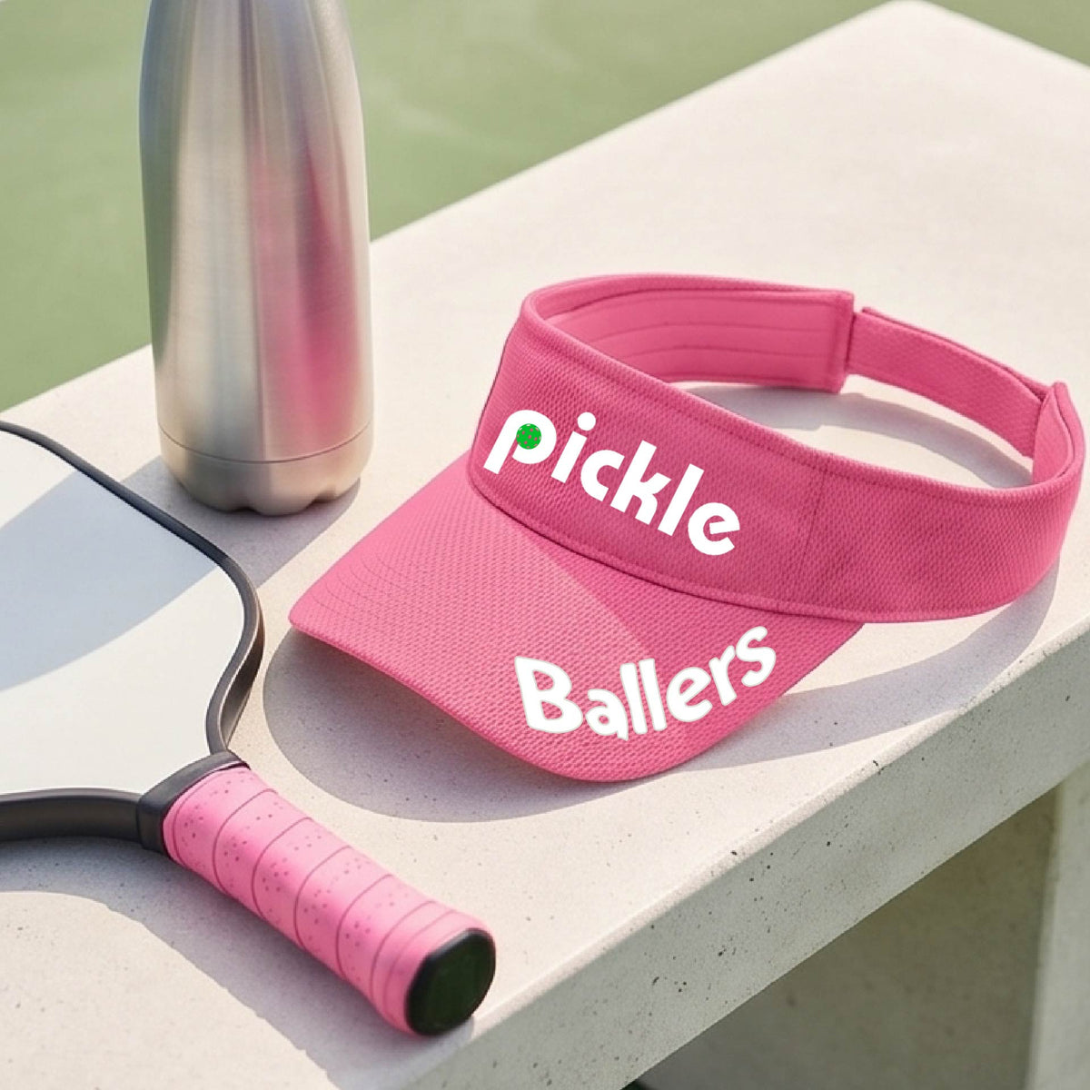 A close up of a pink visor with a pickleball graphic of 'pickle' and 'Ballers' printed in white on the front and brim.