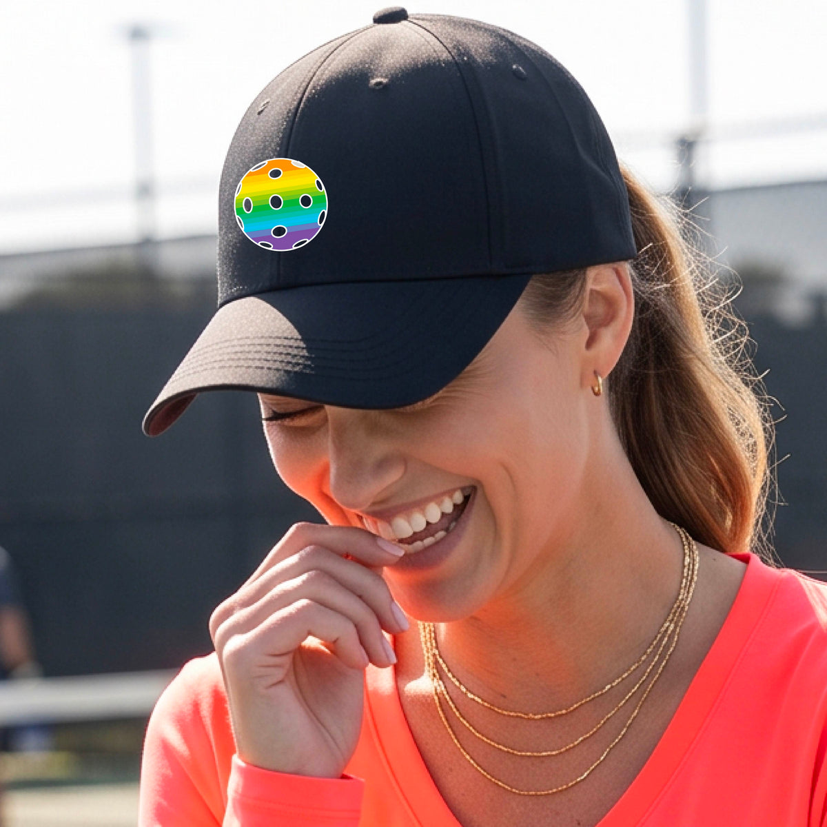 A woman wearing a black baseball cap with a colorful rainbow pickleball on the front, paired with a bright coral-pink top and layered gold necklaces, captured in an outdoor setting.