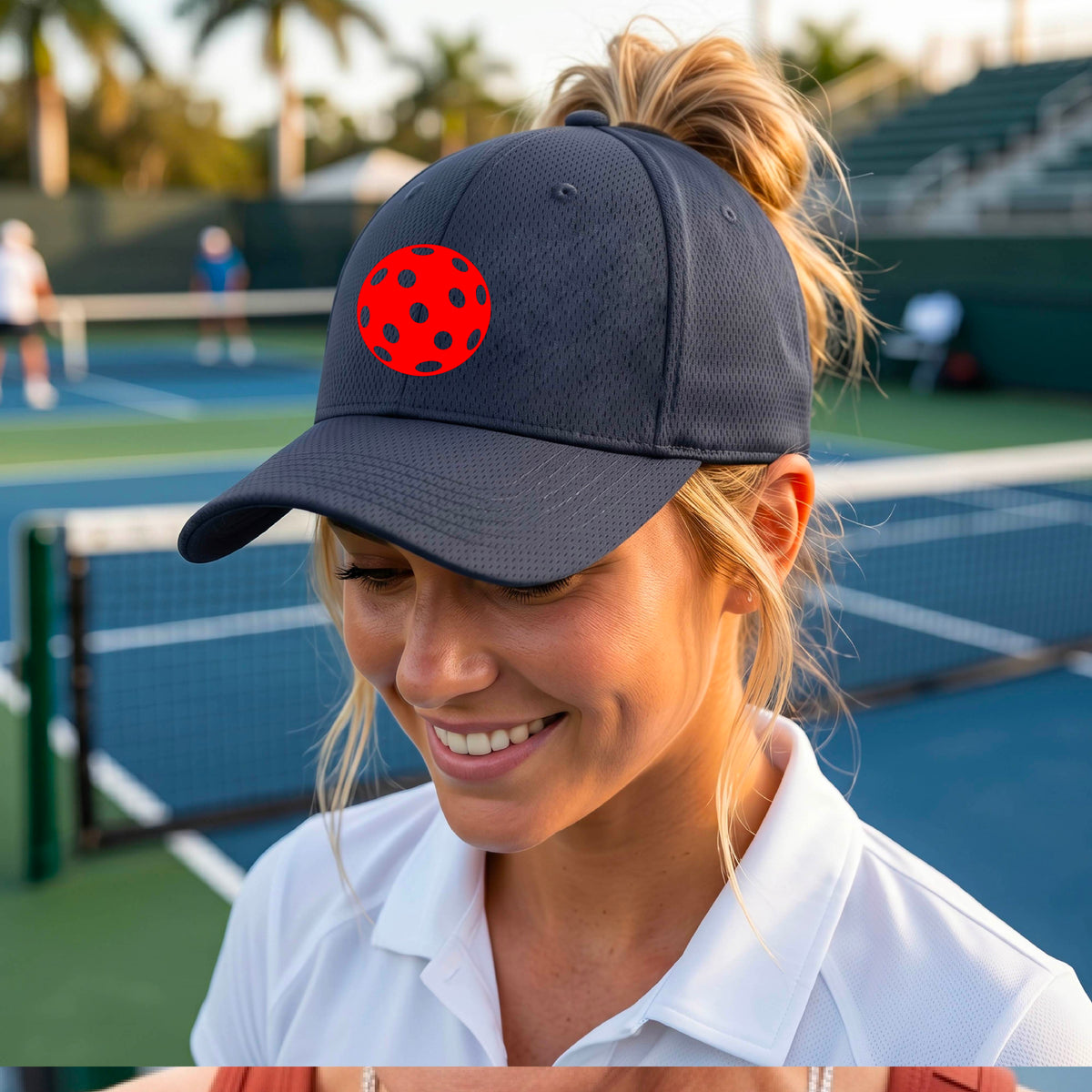 A woman on a pickleball court wearing a dark navy blue visor with a red pickleball The background shows a blue pickleball  court with other players and palm trees in the distance.