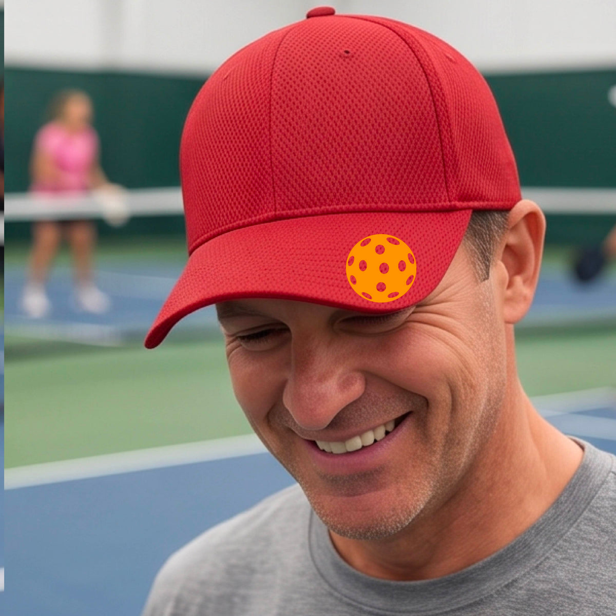 A smiling man wearing a red baseball cap with a yellow-orange pickleball graphic on the front, photographed on a tennis court with another player blurred in the background.
