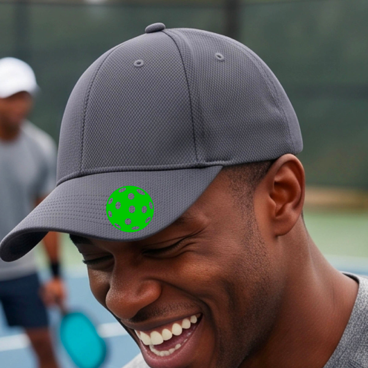 A man smiling on a pickleball court wearing a gray baseball cap with a bright green pickleball graphic on the brim.