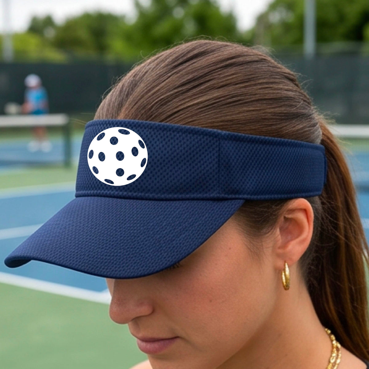 A close-up of a person  wearing a navy blue visor with a pickleball graphic, set against a blurred pickleball court background.