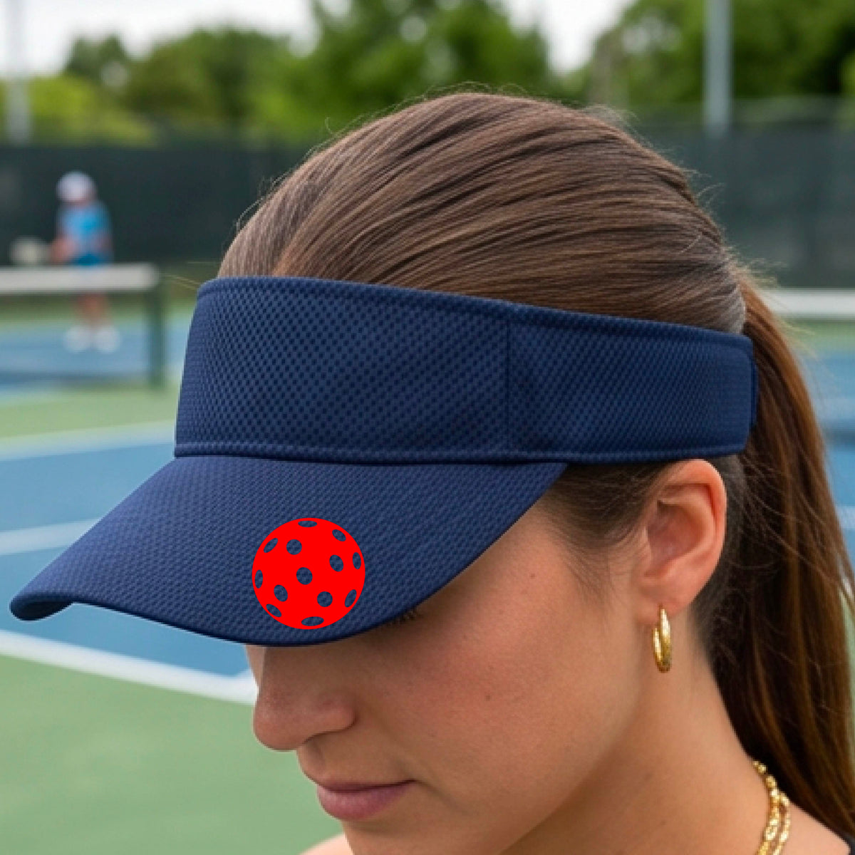 A close-up of a person  wearing a navy blue visor with a pickleball graphic, set against a blurred pickleball court background.