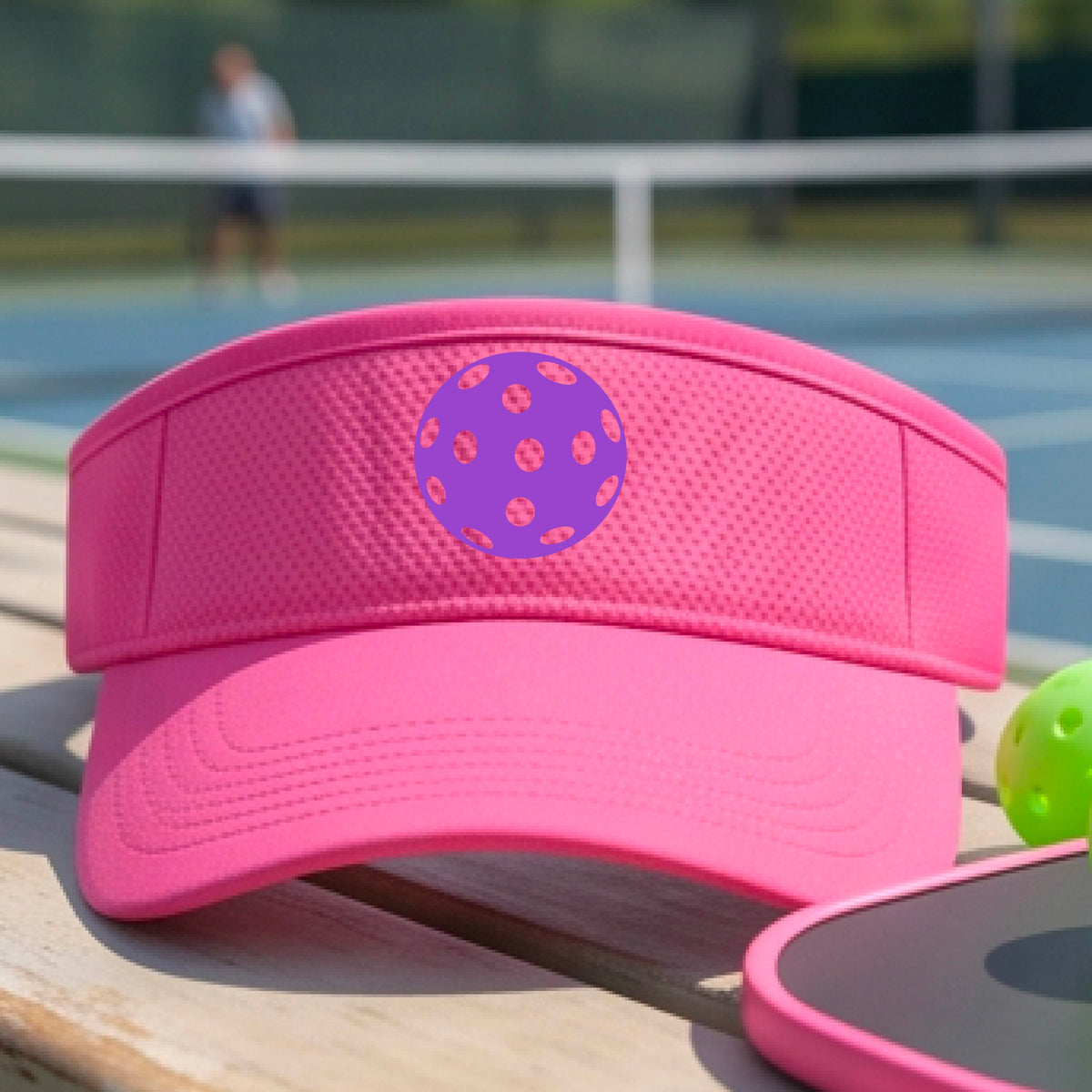 A close-up of a person  wearing a pink visor with a pickleball graphic, set against a blurred pickleball court background.
