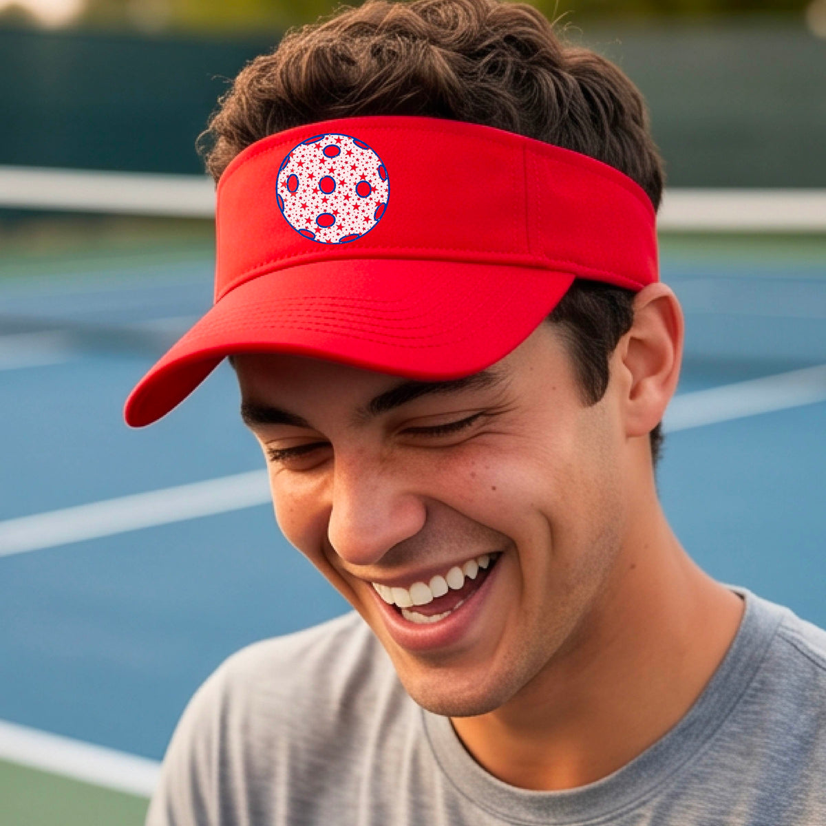 A close-up of a person  wearing a red visor with a pickleball graphic, set against a blurred pickleball court background.
