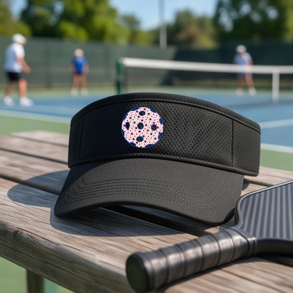 A close-up of a person  wearing a black visor with a pickleball graphic, set against a blurred pickleball court background.