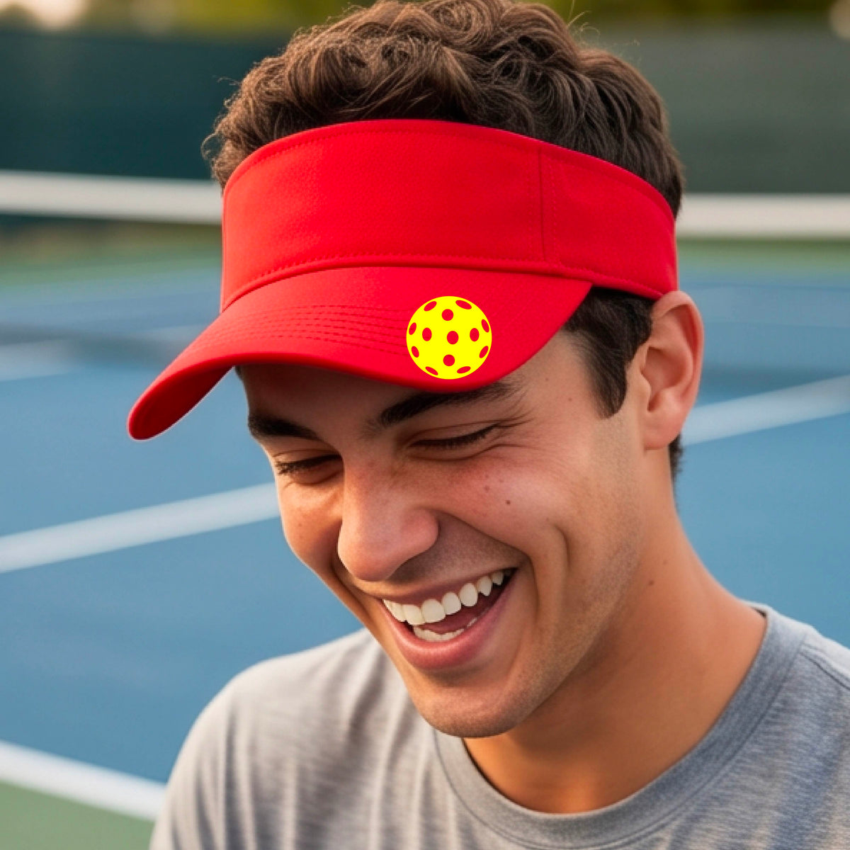 A close-up of a person  wearing a red visor with a pickleball graphic, set against a blurred pickleball court background.