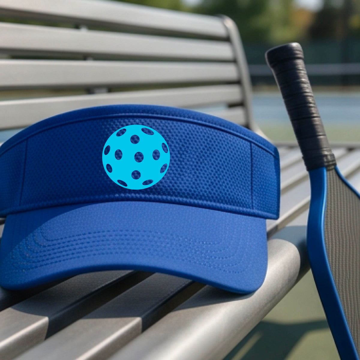 A close-up of a person wearing a royal blue visor with a pickleball graphic, set against a blurred pickleball court background.