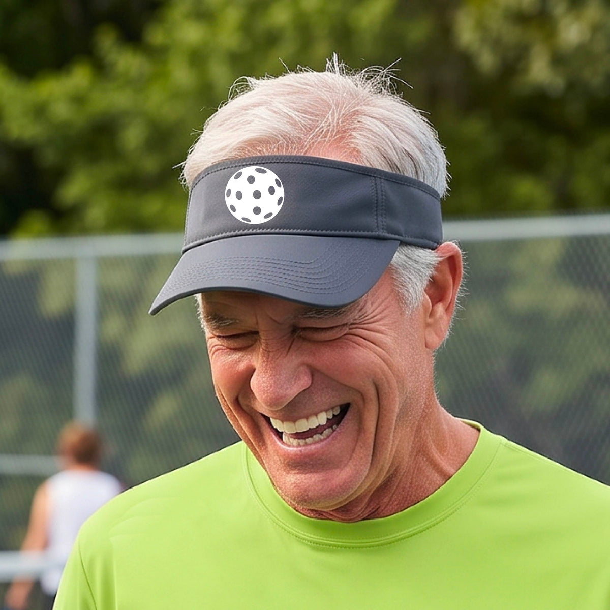 A close-up of a person  wearing a gray visor with a pickleball graphic, set against a blurred pickleball court background.
