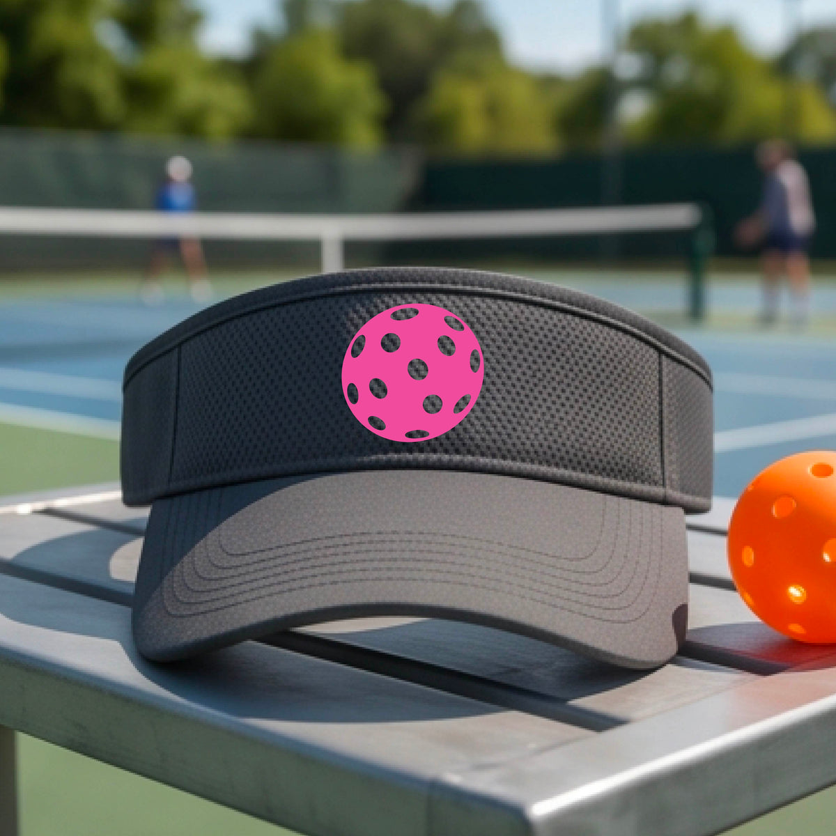 A close-up of a person  wearing a dark gray visor with a pickleball graphic, set against a blurred pickleball court background.