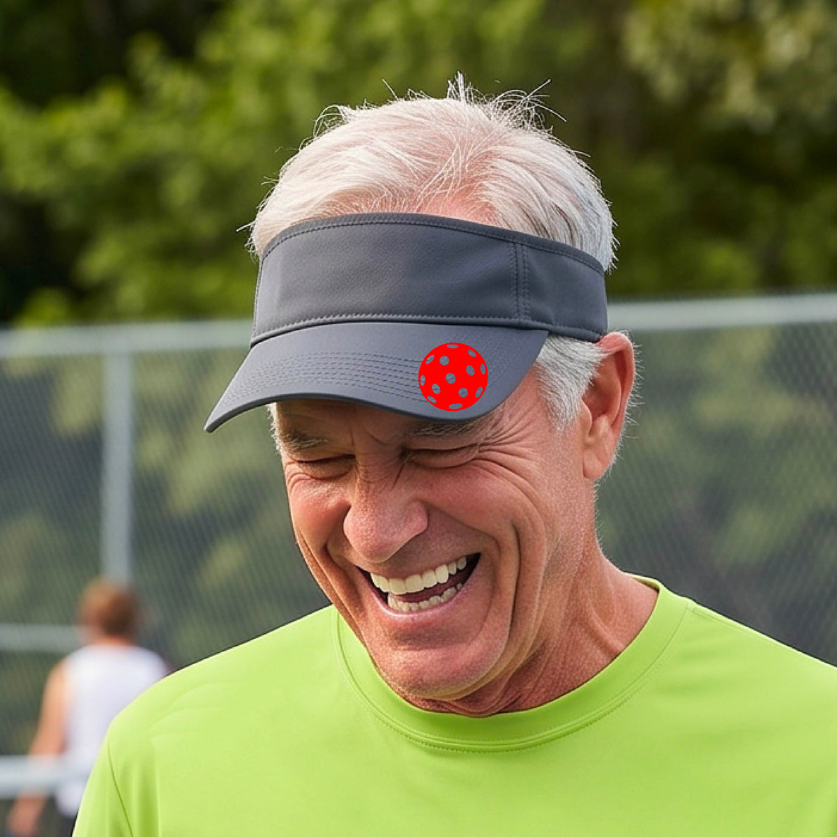 A close-up of a person  wearing a gray visor with a pickleball graphic, set against a blurred pickleball court background.