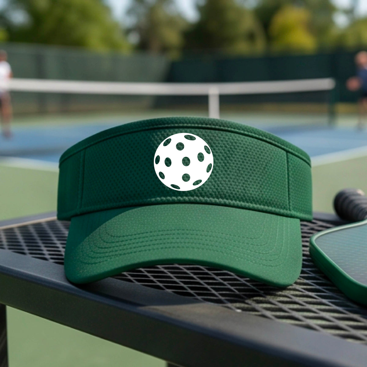 A close-up of a person wearing a green visor with a pickleball graphic, set against a blurred pickleball court background.