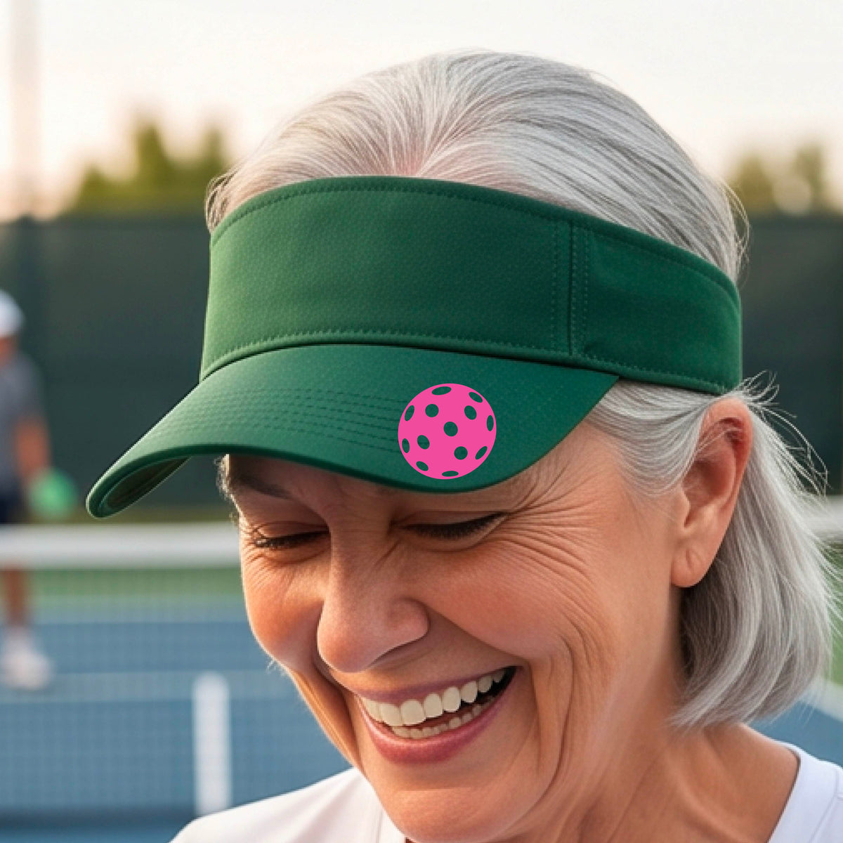 A close-up of a person  wearing a green visor with a pickleball graphic, set against a blurred pickleball court background.