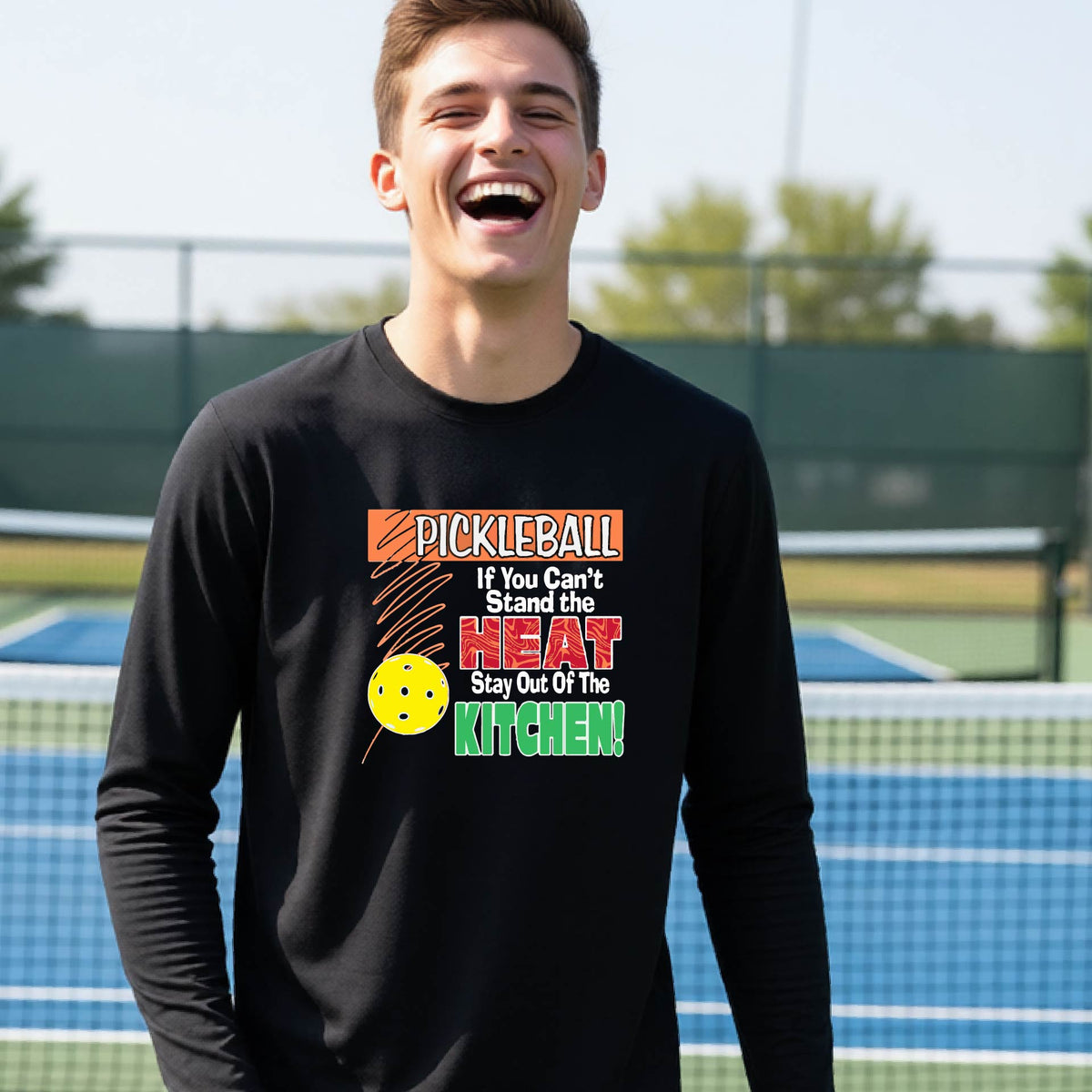 A close up of a young person wearing a black long-sleeve t-shirt with a colorful pickleball graphic and text on the front "IF YOU CAN'T STAND THE HEAT STAY OUT OF THE KITCHEN."