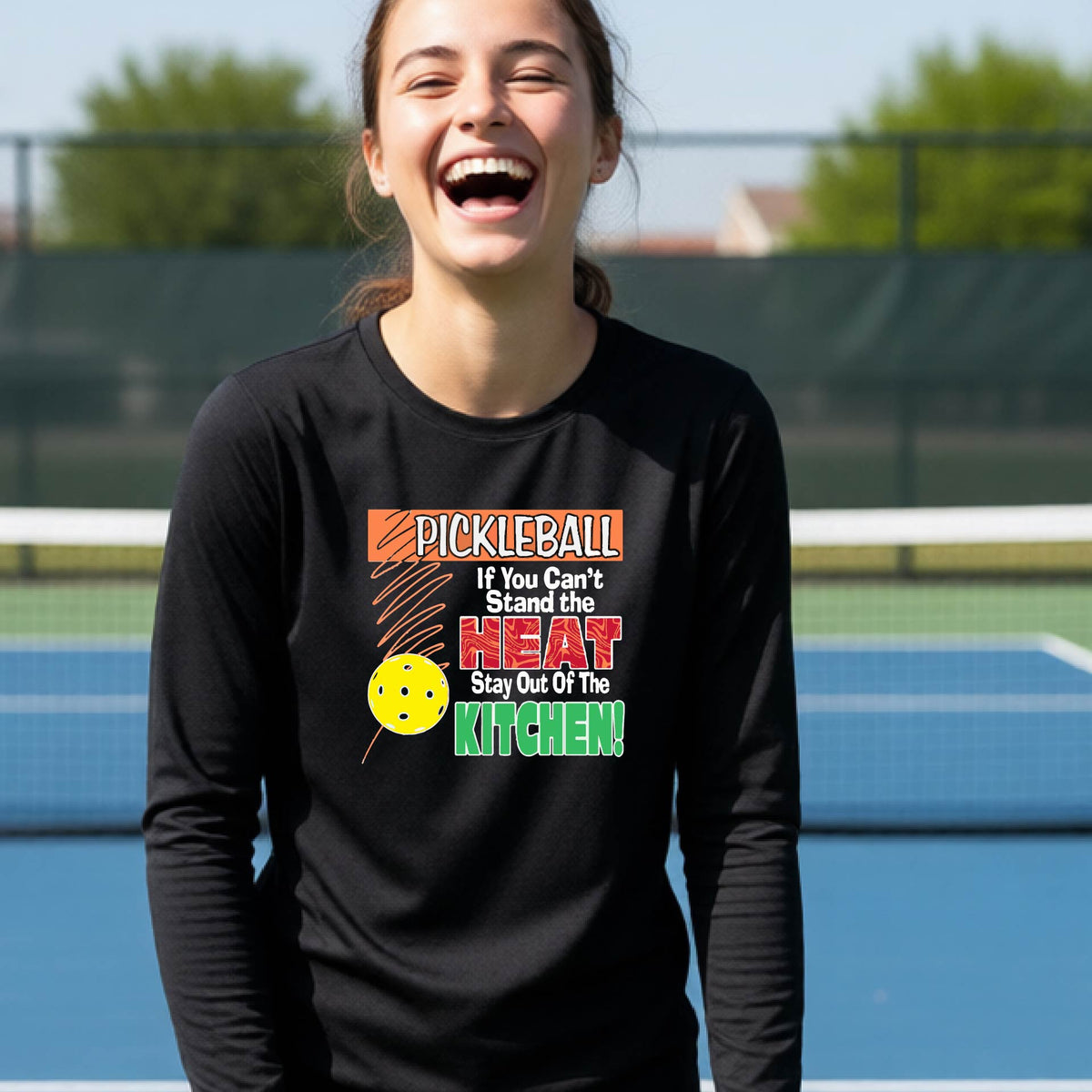 A close up of a young person wearing a black long-sleeve t-shirt with a colorful pickleball graphic and text on the front "IF YOU CAN'T STAND THE HEAT STAY OUT OF THE KITCHEN."
