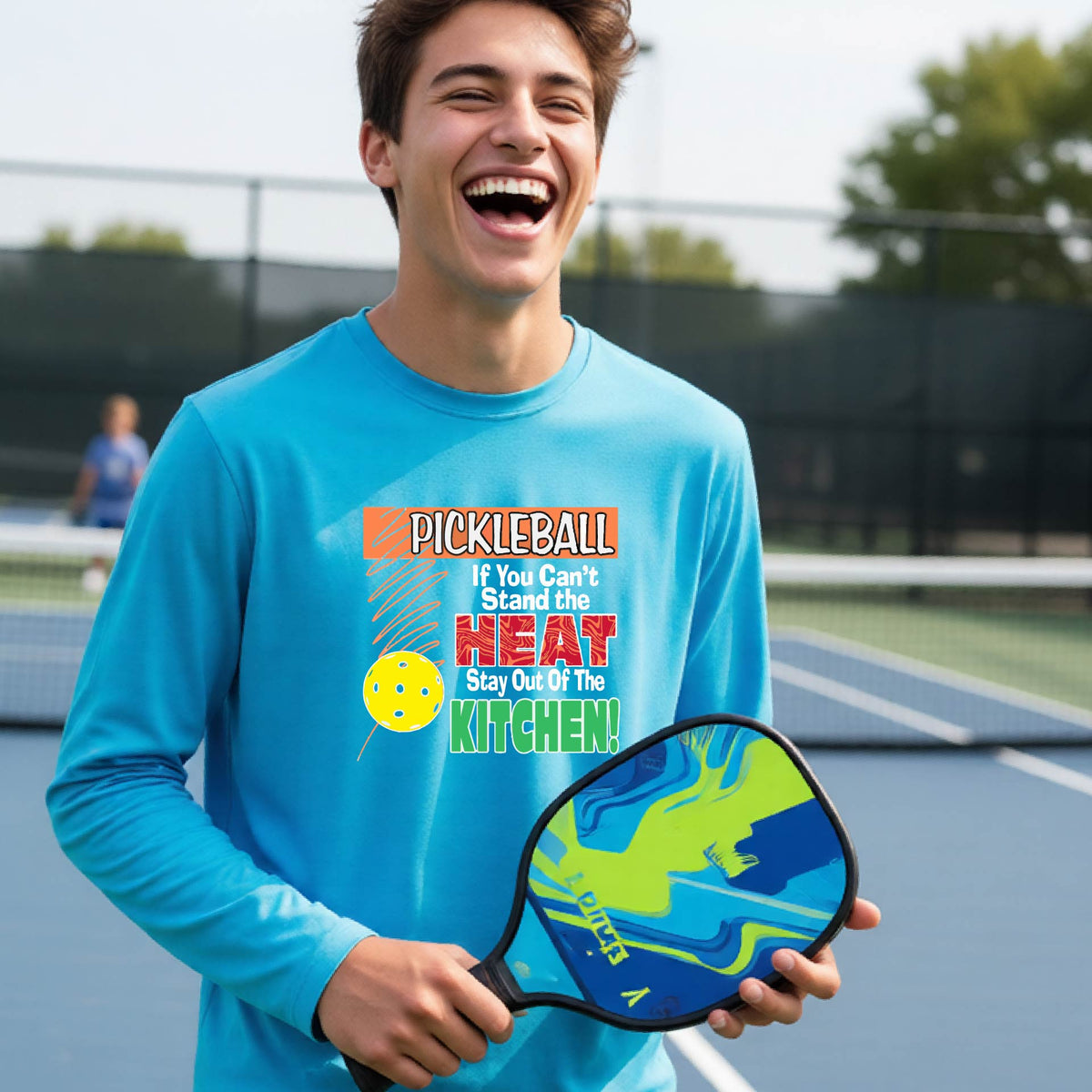 A close up of a young person wearing a light blue long-sleeve t-shirt with a colorful pickleball graphic and text on the front "IF YOU CAN'T STAND THE HEAT STAY OUT OF THE KITCHEN."