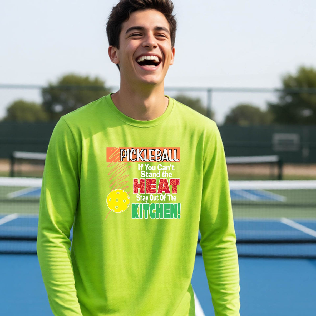A close up of a young person wearing a lime green long-sleeve t-shirt with a colorful pickleball graphic and text on the front "IF YOU CAN'T STAND THE HEAT STAY OUT OF THE KITCHEN."