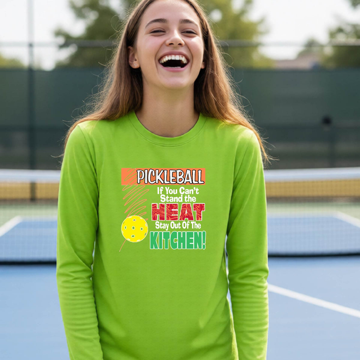 A close up of a young person wearing a lime green long-sleeve t-shirt with a colorful pickleball graphic and text on the front "IF YOU CAN'T STAND THE HEAT STAY OUT OF THE KITCHEN."
