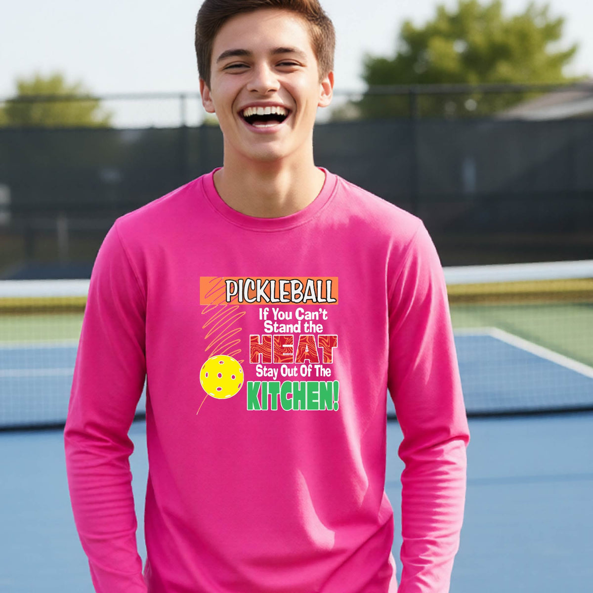 A close up of a young person wearing a bright pink long-sleeve t-shirt with a colorful pickleball graphic and text on the front "IF YOU CAN'T STAND THE HEAT STAY OUT OF THE KITCHEN."