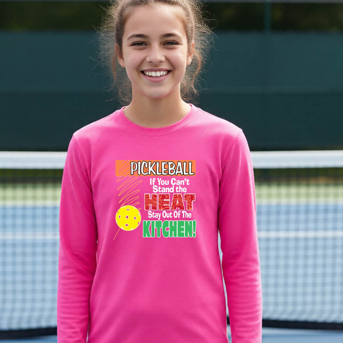 A close up of a young person wearing a bright pink long-sleeve t-shirt with a colorful pickleball graphic and text on the front "IF YOU CAN'T STAND THE HEAT STAY OUT OF THE KITCHEN."