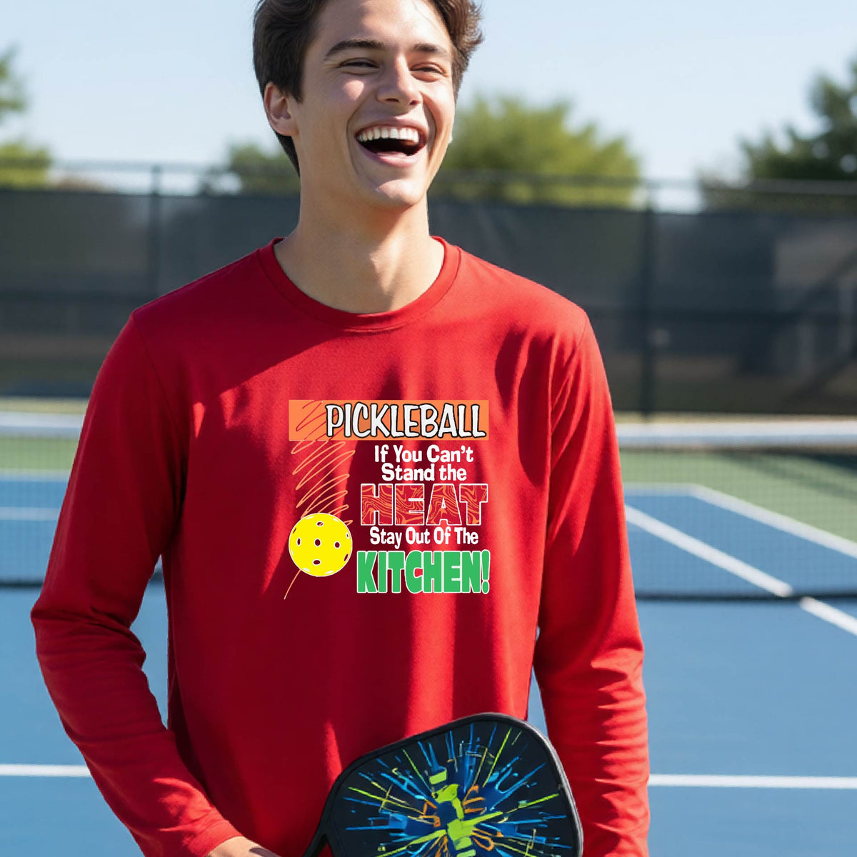 A close up of a young person wearing a red long-sleeve t-shirt with a colorful pickleball graphic and text on the front "IF YOU CAN'T STAND THE HEAT STAY OUT OF THE KITCHEN."