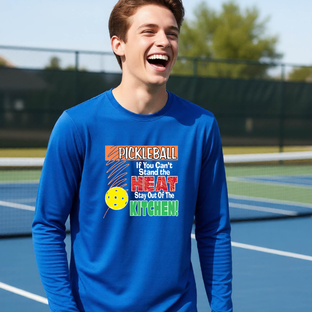A close up of a young person wearing a royal blue long-sleeve t-shirt with a colorful pickleball graphic and text on the front "IF YOU CAN'T STAND THE HEAT STAY OUT OF THE KITCHEN."