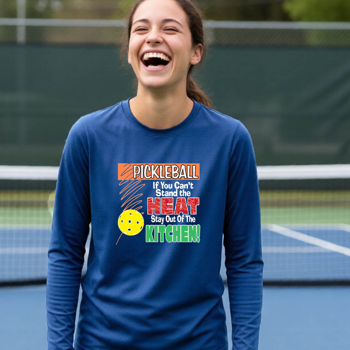 A close up of a young person wearing a royal blue long-sleeve t-shirt with a colorful pickleball graphic and text on the front "IF YOU CAN'T STAND THE HEAT STAY OUT OF THE KITCHEN."