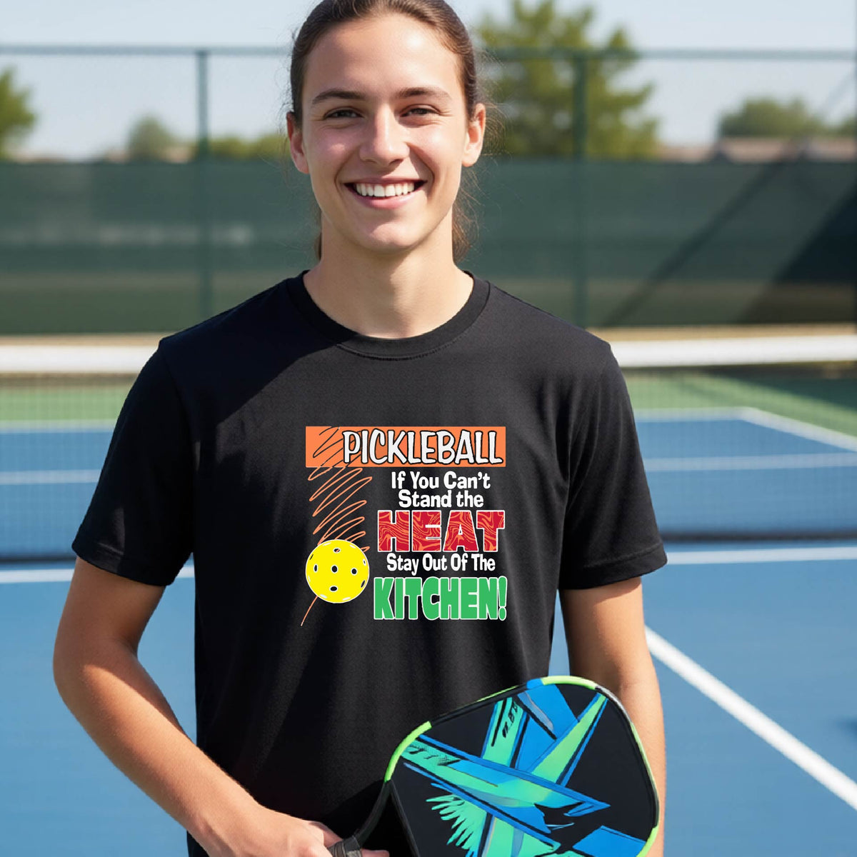 A young person laughing on an outdoor pickleall court wearing a black graphic t-shirt with a pickleball-themed slogan ":PICKLEBALL IF YOU CAN'T STAND THE HEAT STAY OUT OF THE KITCHEN"