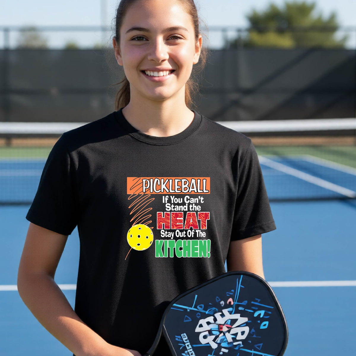 A young person laughing on an outdoor pickleall court wearing a black graphic t-shirt with a pickleball-themed slogan ":PICKLEBALL IF YOU CAN'T STAND THE HEAT STAY OUT OF THE KITCHEN"