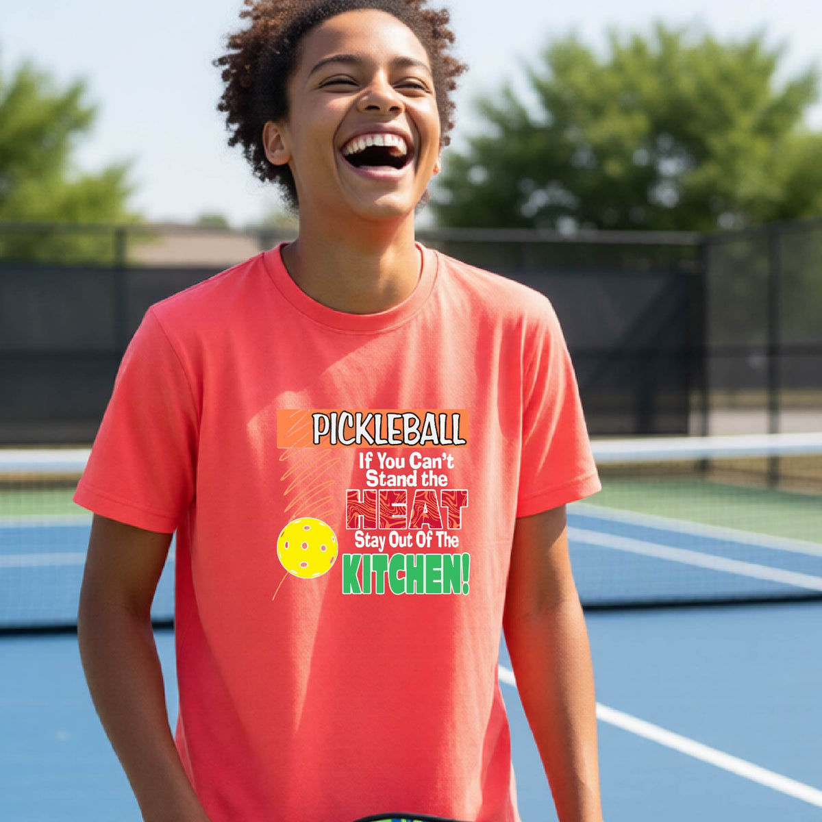 A young person laughing on an outdoor pickleall court wearing a coral pink graphic t-shirt with a pickleball-themed slogan ":PICKLEBALL IF YOU CAN'T STAND THE HEAT STAY OUT OF THE KITCHEN"