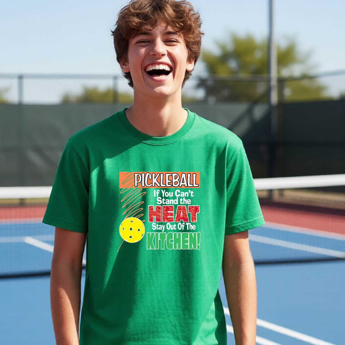 A young person laughing on an outdoor pickleall court wearing a green graphic t-shirt with a pickleball-themed slogan ":PICKLEBALL IF YOU CAN'T STAND THE HEAT STAY OUT OF THE KITCHEN"