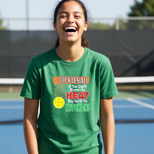 A young person laughing on an outdoor pickleall court wearing a green graphic t-shirt with a pickleball-themed slogan ":PICKLEBALL IF YOU CAN'T STAND THE HEAT STAY OUT OF THE KITCHEN"