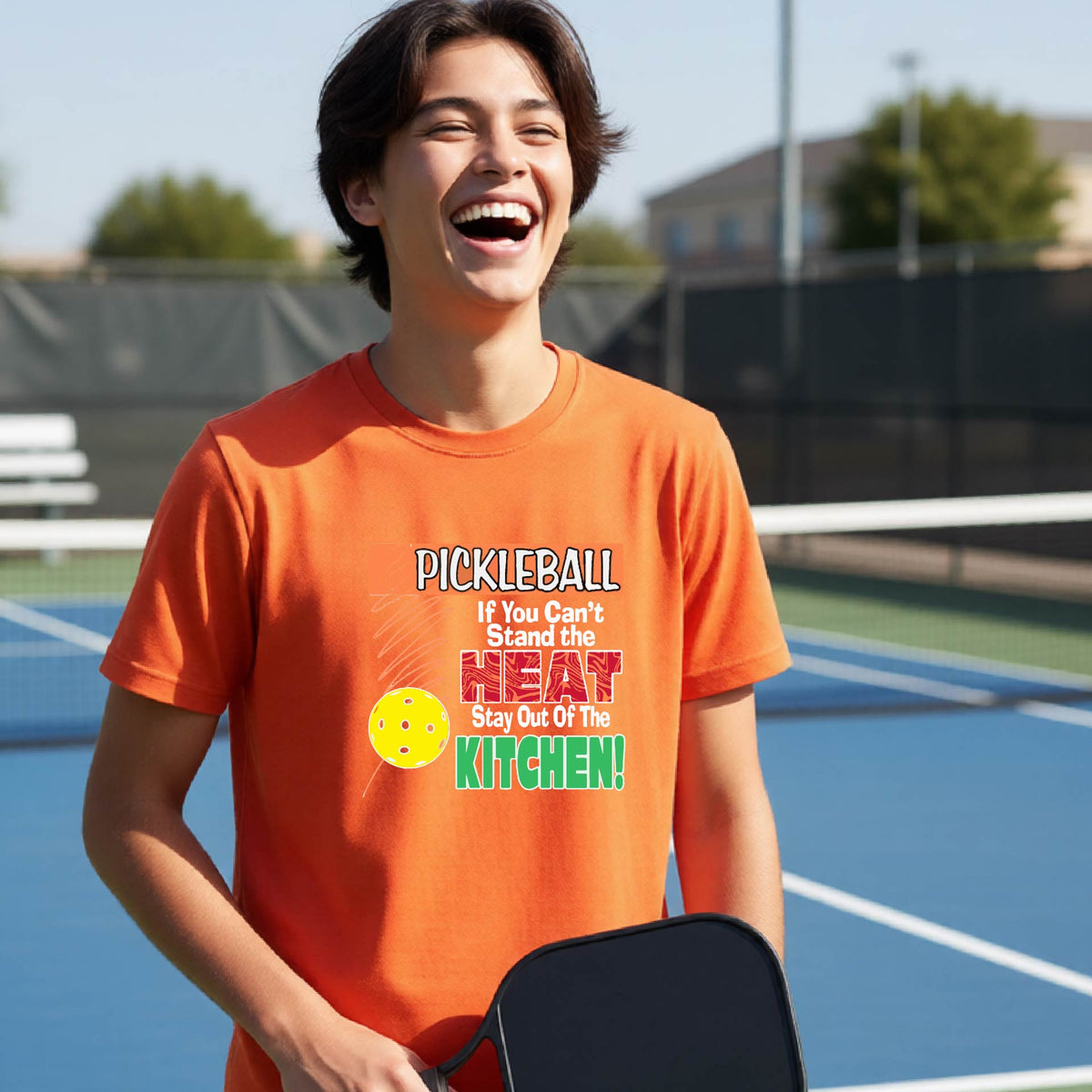A young person laughing on an outdoor pickleall court wearing an orange graphic t-shirt with a pickleball-themed slogan ":PICKLEBALL IF YOU CAN'T STAND THE HEAT STAY OUT OF THE KITCHEN"