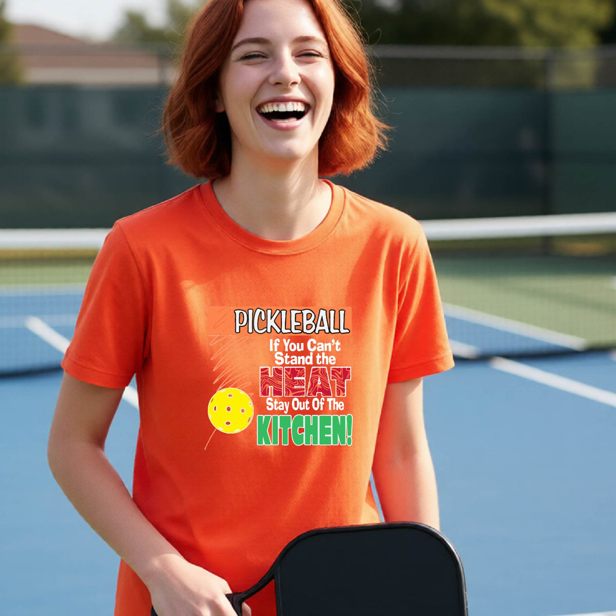 A young person laughing on an outdoor pickleall court wearing an orange graphic t-shirt with a pickleball-themed slogan ":PICKLEBALL IF YOU CAN'T STAND THE HEAT STAY OUT OF THE KITCHEN"