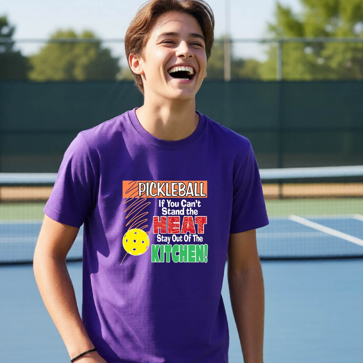 A young person laughing on an outdoor pickleall court wearing a purple graphic t-shirt with a pickleball-themed slogan ":PICKLEBALL IF YOU CAN'T STAND THE HEAT STAY OUT OF THE KITCHEN"
