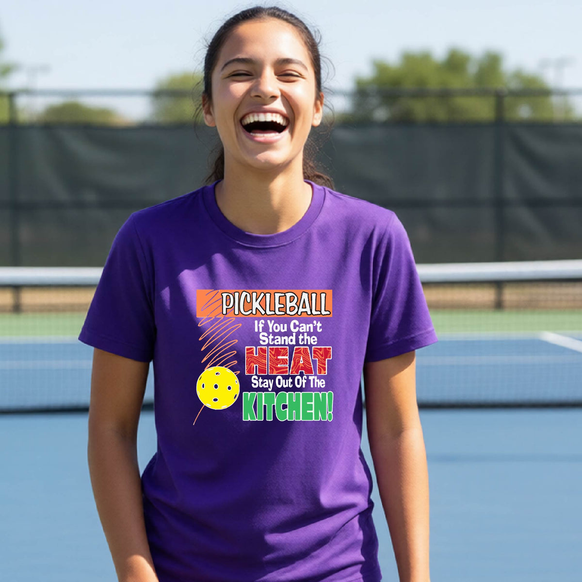 A young person laughing on an outdoor pickleall court wearing a purple graphic t-shirt with a pickleball-themed slogan ":PICKLEBALL IF YOU CAN'T STAND THE HEAT STAY OUT OF THE KITCHEN"