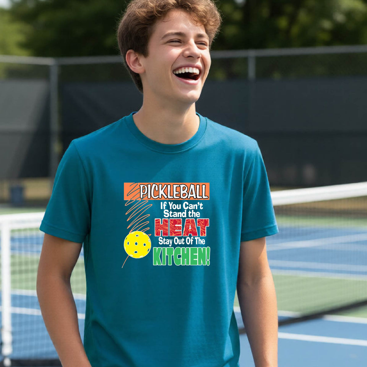 A young person laughing on an outdoor pickleall court wearing a  teal graphic t-shirt with a pickleball-themed slogan ":PICKLEBALL IF YOU CAN'T STAND THE HEAT STAY OUT OF THE KITCHEN"
