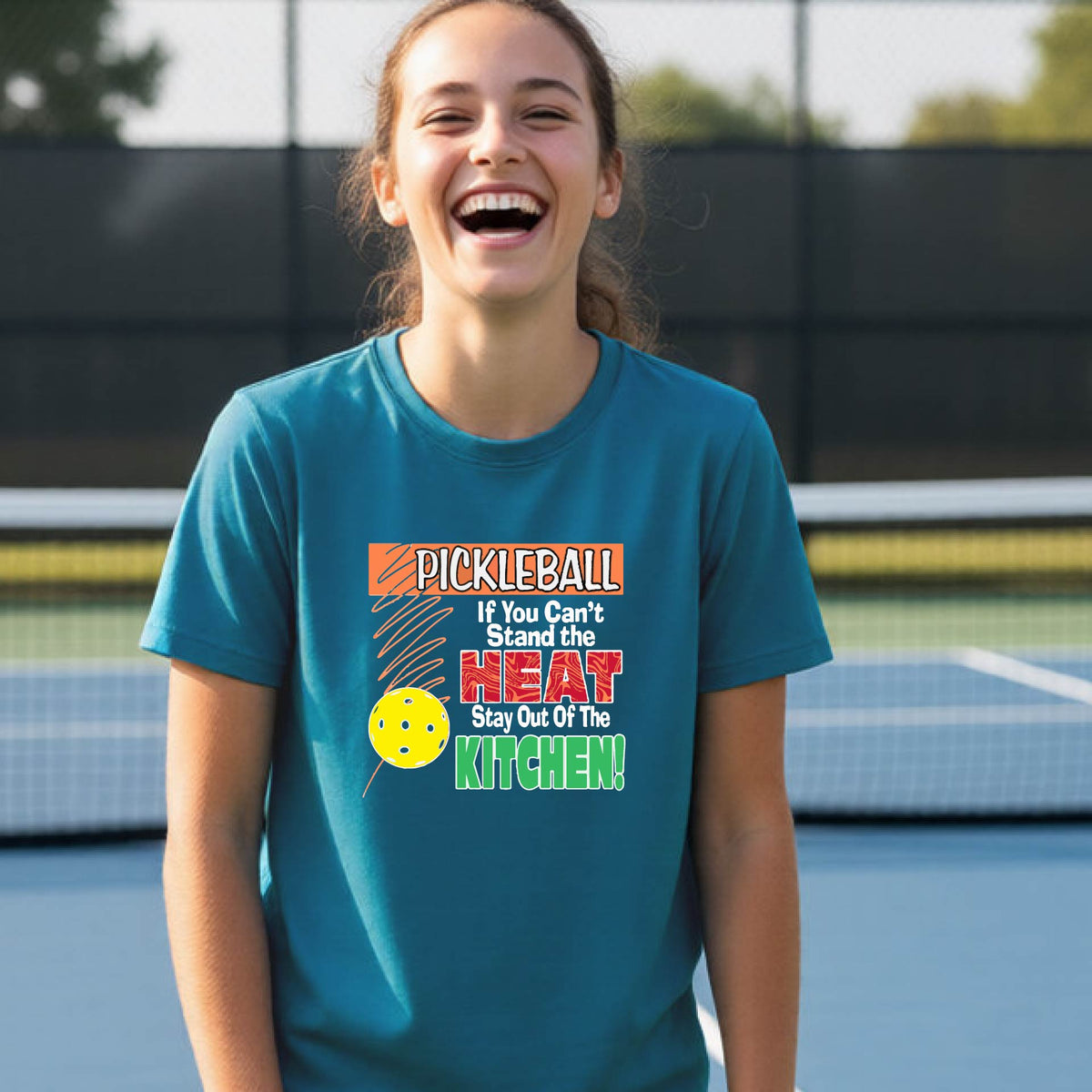 A young person laughing on an outdoor pickleall court wearing a teal graphic t-shirt with a pickleball-themed slogan ":PICKLEBALL IF YOU CAN'T STAND THE HEAT STAY OUT OF THE KITCHEN"