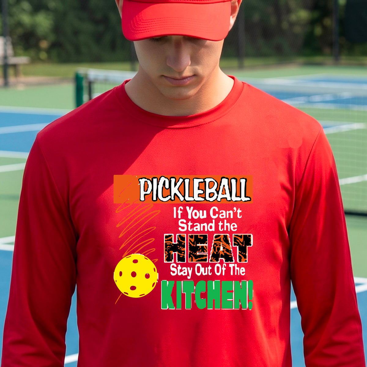 A smiling man wearing a red long-sleeve t-shirt with a colorful pickleball graphic text “If You Can't Stand The Heat Stay Out Of The Kitchen" on the chest, standing on an outdoor pickleball court with mountains in the background.
