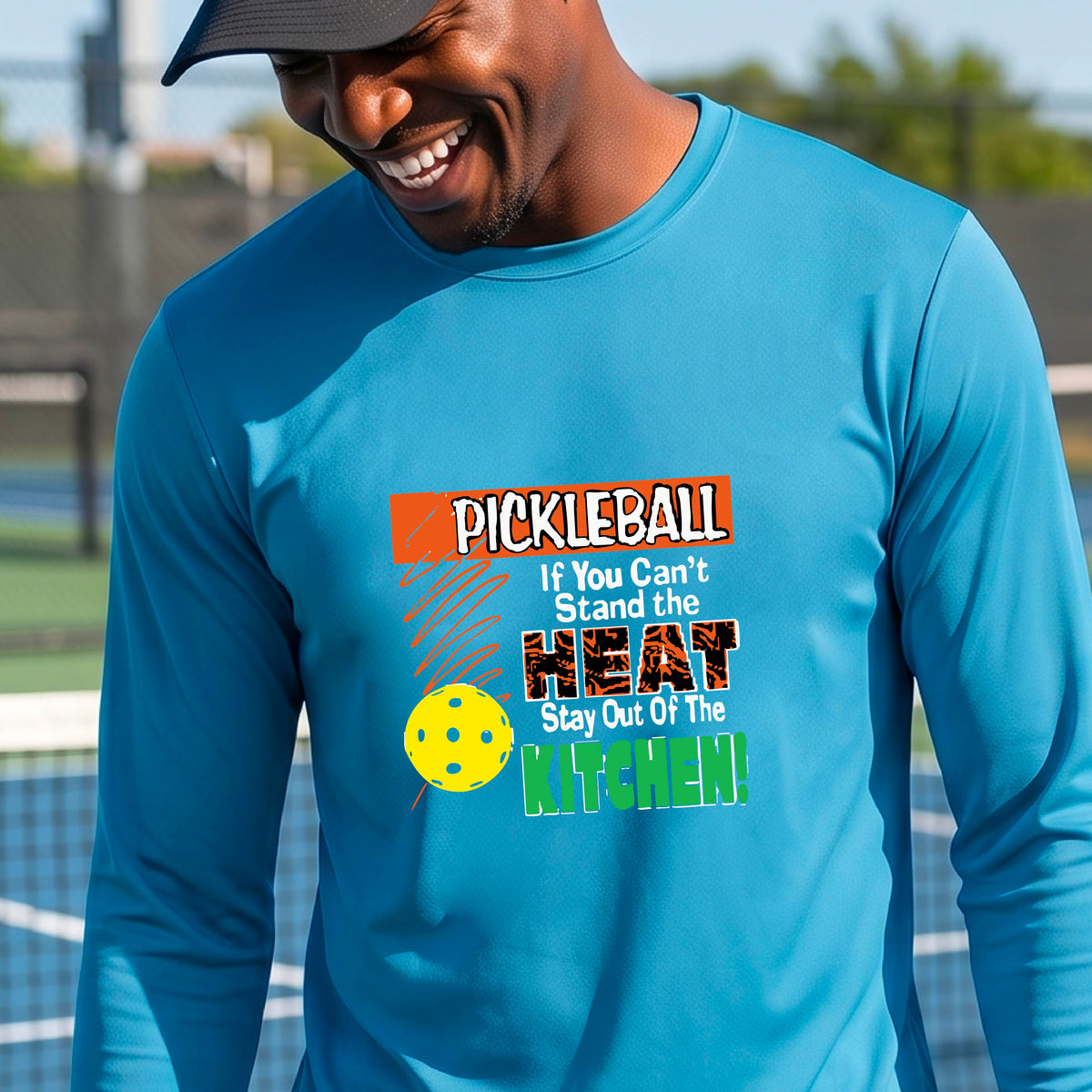 A smiling man wearing a bright blue long-sleeve t-shirt with a colorful pickleball graphic text “If You Can't Stand The Heat Stay Out Of The Kitchen" on the chest, standing on an outdoor pickleball court with mountains in the background.