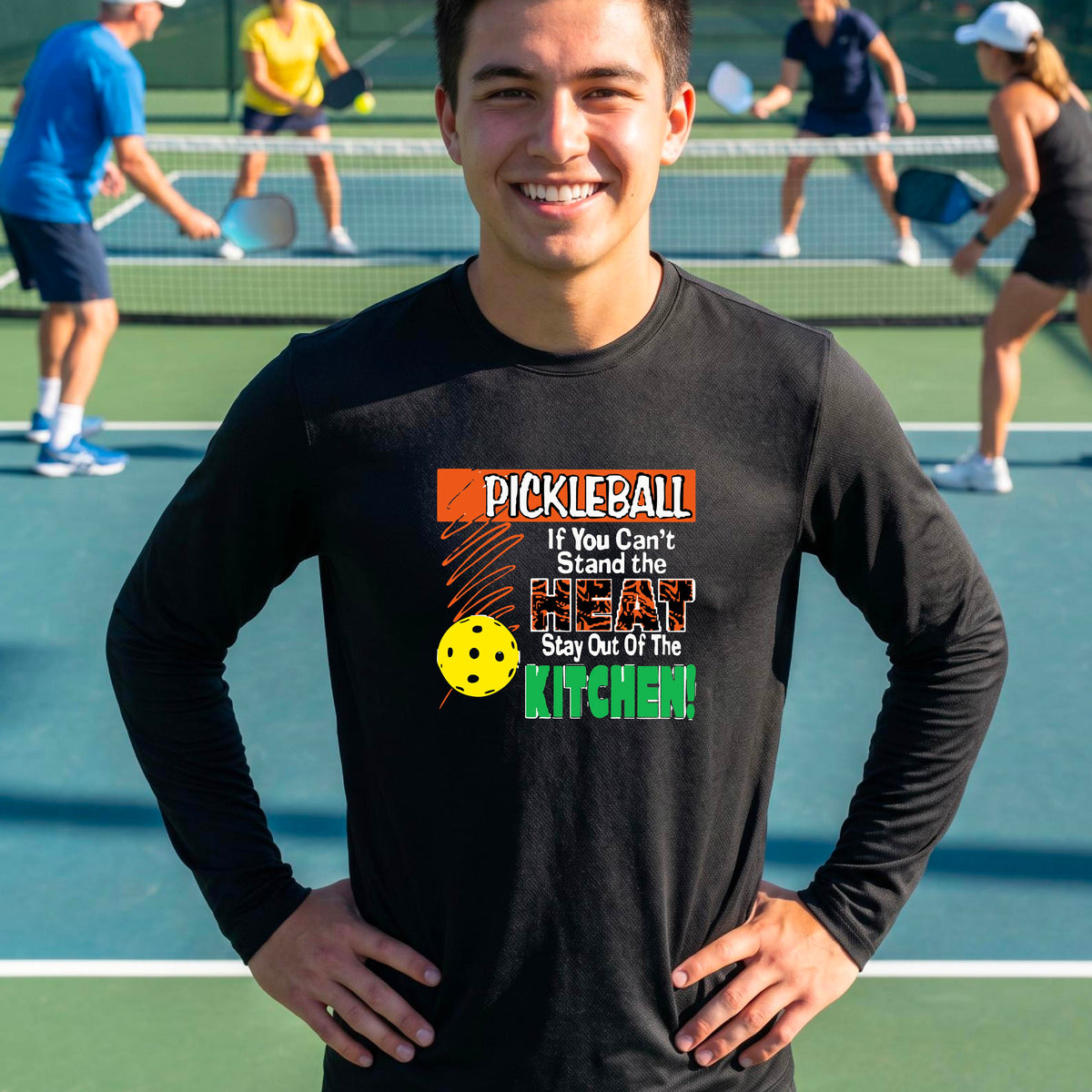 A smiling man wearing a black long-sleeve t-shirt with a colorful pickleball graphic text “If You Can't Stand The Heat Stay Out Of The Kitchen" on the chest, standing on an outdoor pickleball court with mountains in the background.