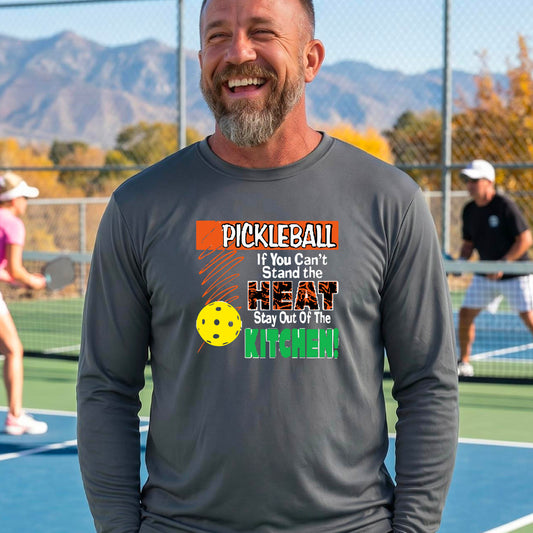 A smiling man wearing a dark gray long-sleeve t-shirt with a colorful pickleball graphic text “If You Can't Stand The Heat Stay Out Of The Kitchen" on the chest, standing on an outdoor pickleball court with mountains in the background.
