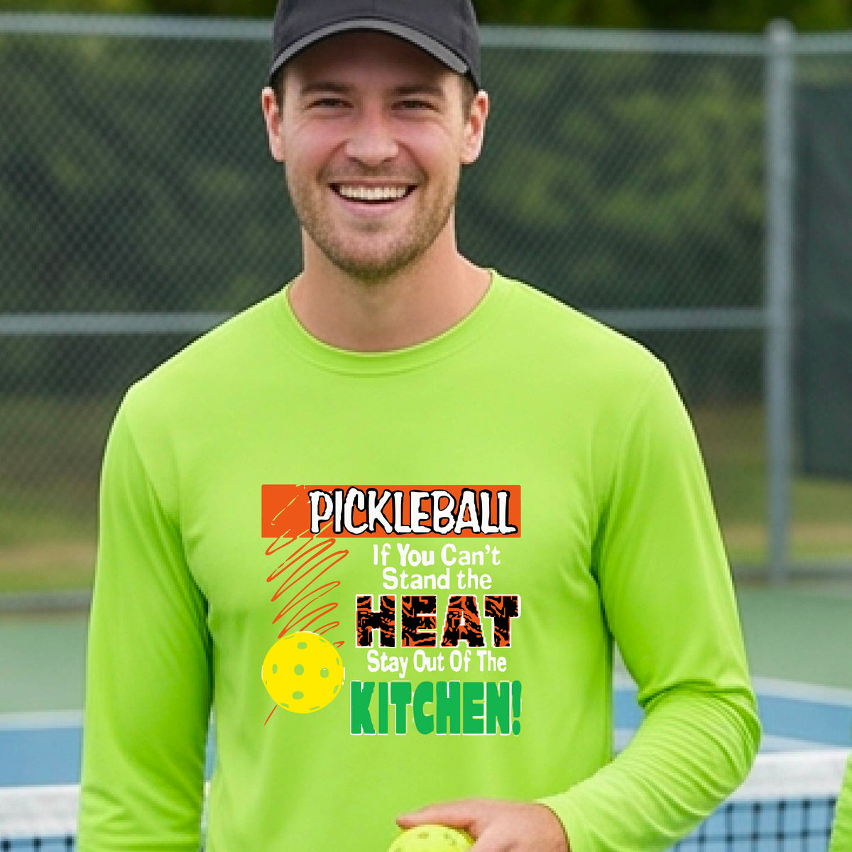 A smiling man wearing a lime green long-sleeve t-shirt with a colorful pickleball graphic text “If You Can't Stand The Heat Stay Out Of The Kitchen" on the chest, standing on an outdoor pickleball court with mountains in the background.