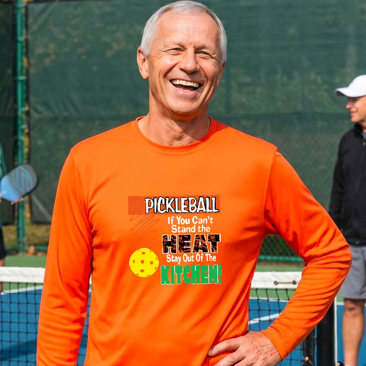 A smiling man wearing an orange long-sleeve t-shirt with a colorful pickleball graphic text “If You Can't Stand The Heat Stay Out Of The Kitchen" on the chest, standing on an outdoor pickleball court with mountains in the background.