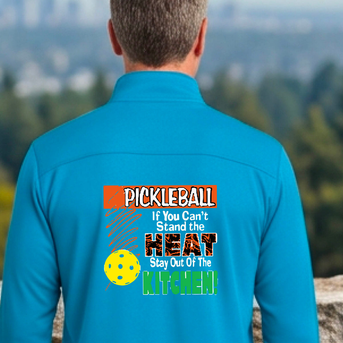 A man viewed from behind wearing a bright blue long-sleeve quarter-zip pullover with a colorful pickleball-themed graphic on the back. The graphic features the word 'PICKLEBALL' in orange, a yellow pickleball, and the text 'If You Can't Stand the HEAT Stay Out Of The KITCHEN!' in white and green.
