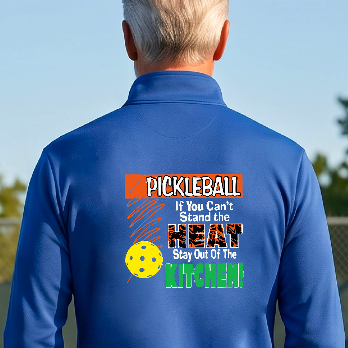 A man viewed from behind wearing a royal blue long-sleeve quarter-zip pullover with a colorful pickleball-themed graphic on the back. The graphic features the word 'PICKLEBALL' in orange, a yellow pickleball, and the text 'If You Can't Stand the HEAT Stay Out Of The KITCHEN!' in white and green.